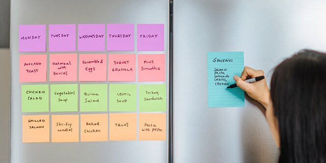 Ein Kühlschrank mit Post-it® Haftnotizen, die den wöchentlichen Essensplan anzeigen, und eine Person, die auf einem linierten Post-it&reg; Note die Einkaufsliste notiert
