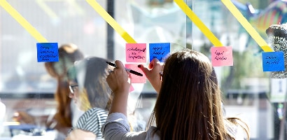 Eine Frau bringt ein Post-it&reg; Super Sticky Note (76&nbsp;mm&nbsp;x&nbsp;76&nbsp;mm) an einer Glaswand an, um Informationen zu Aufgaben oder Projekten mit anderen Teammitgliedern zu teilen.